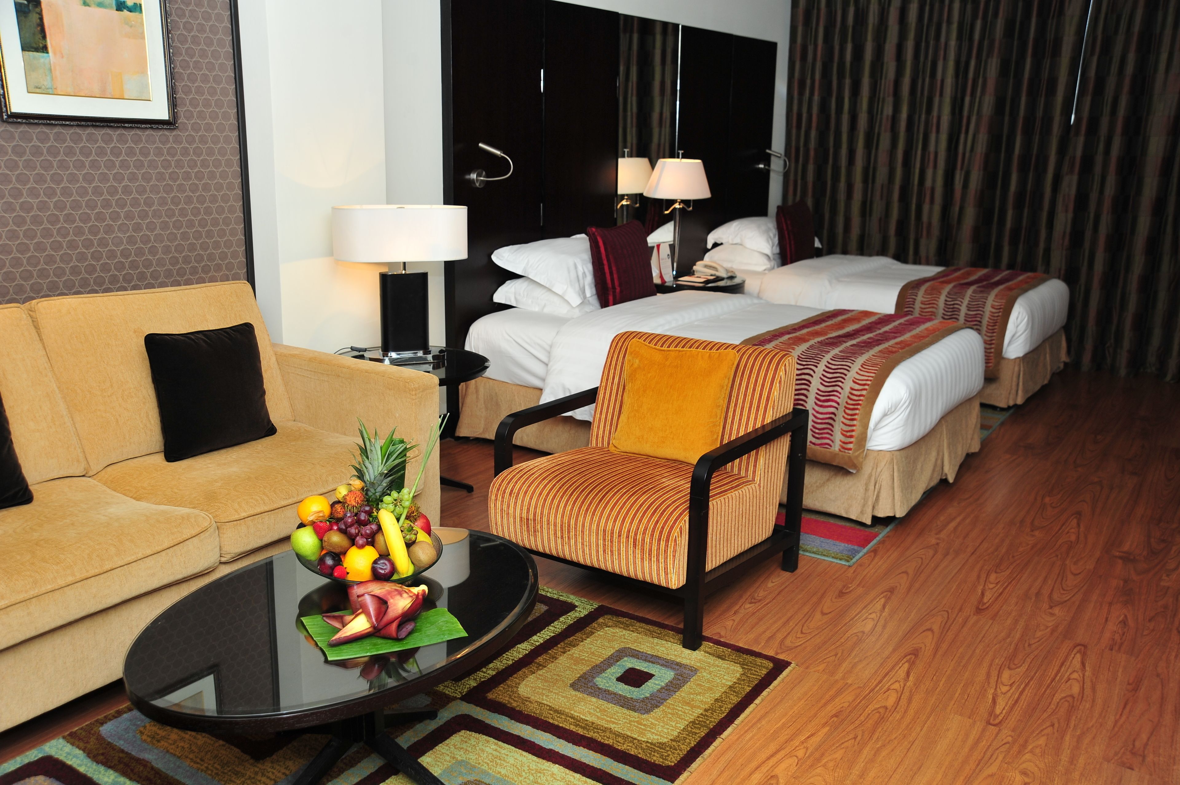 A hotel room with a sofa, two single beds, a chair, a coffee table with fruit, and a lamp.