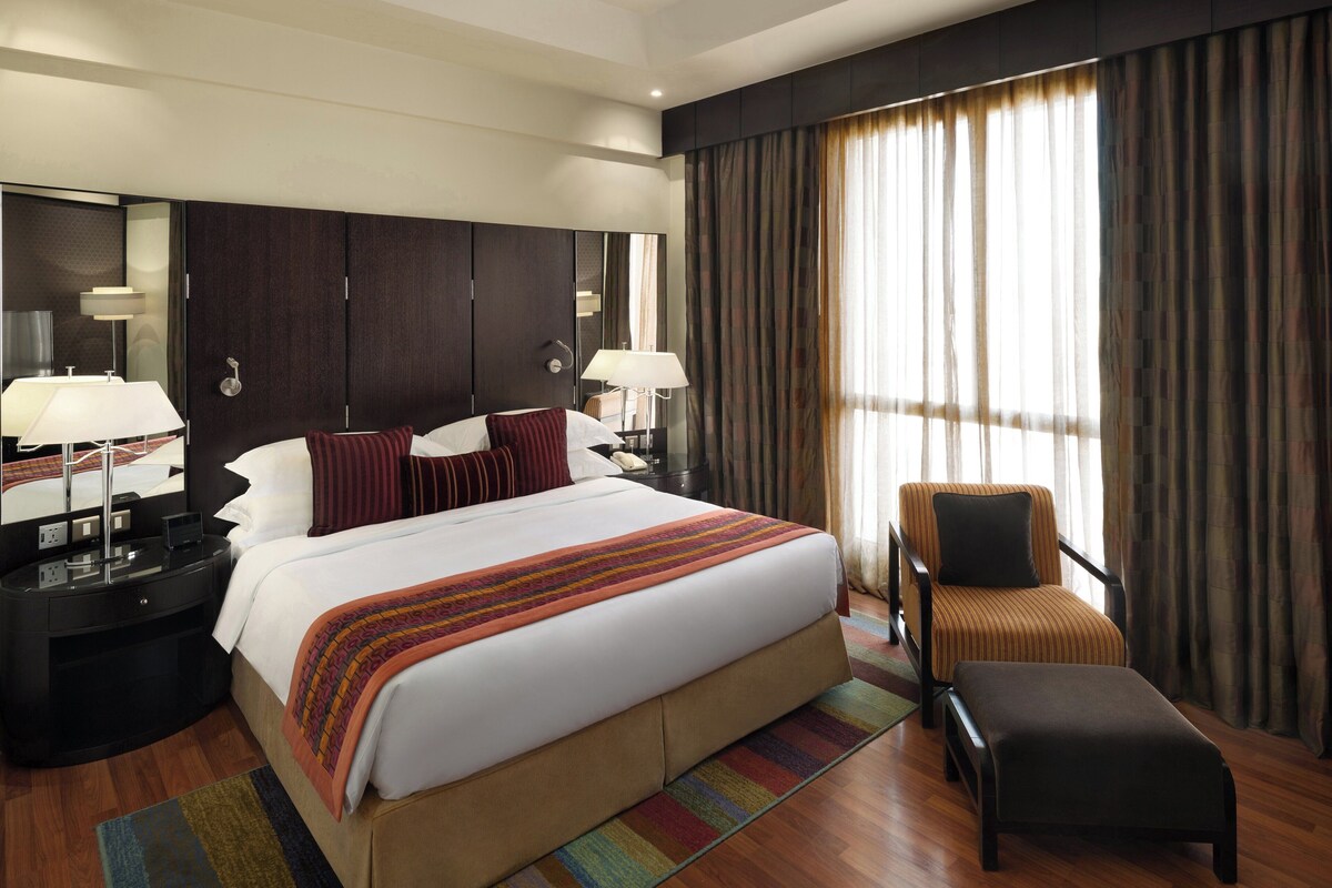 A hotel room with a large bed, bedside tables with lamps, a chair, a desk, and a large window with curtains.