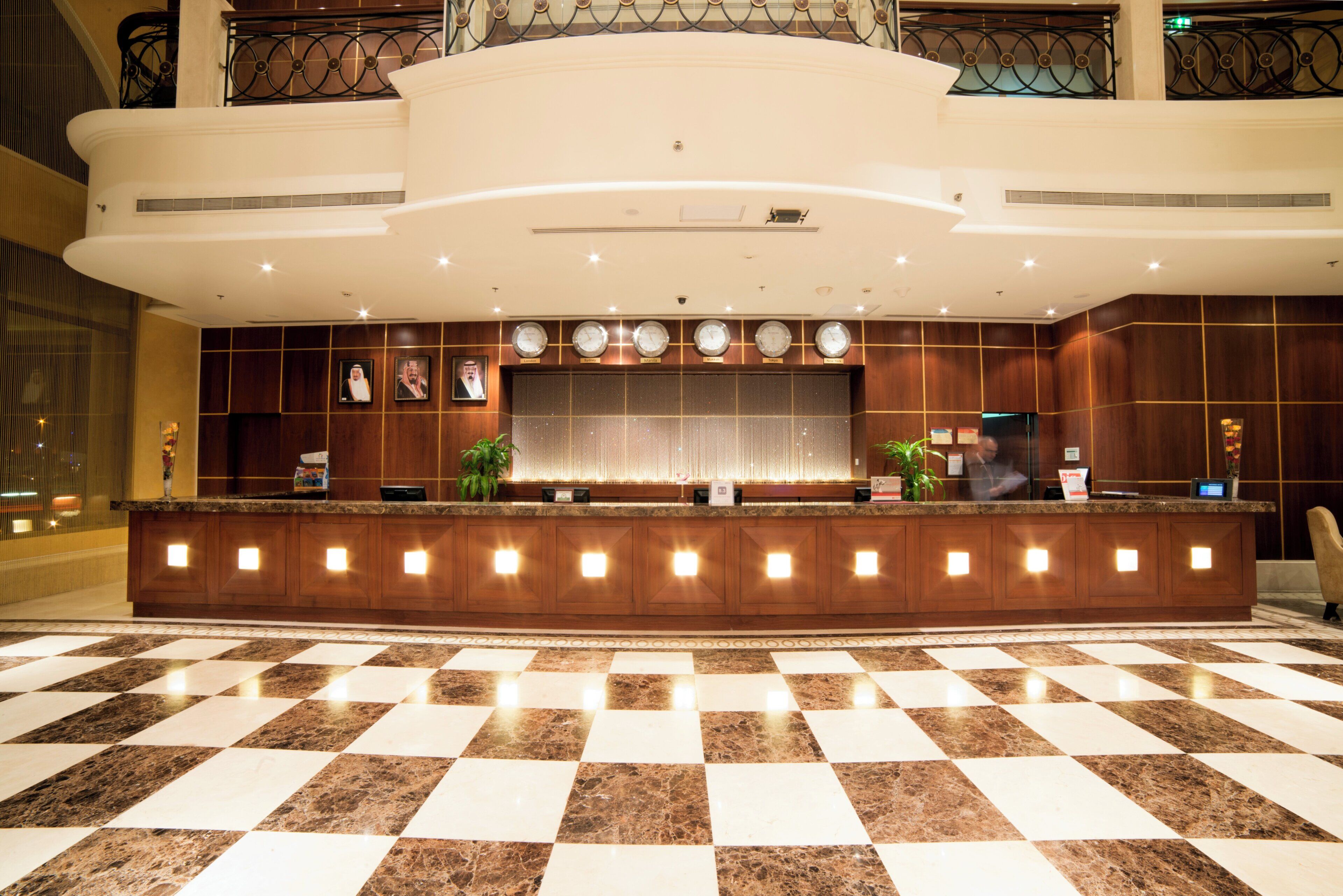 Ein Hotel-Empfangsbereich mit Schachbrettmuster-Boden, einer Rezeption und Uhren, die verschiedene Zeitzonen anzeigen.
