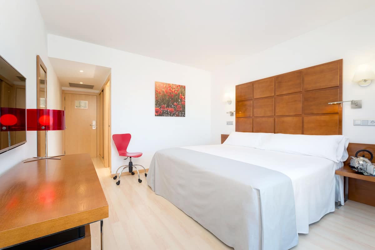 Habitación de hotel con cama, escritorio, silla, cabecero de madera y lámpara fijada a la pared.