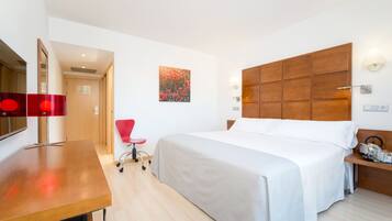 A hotel room with a bed, a desk, a chair, a wooden headboard, and a wall-mounted lamp.