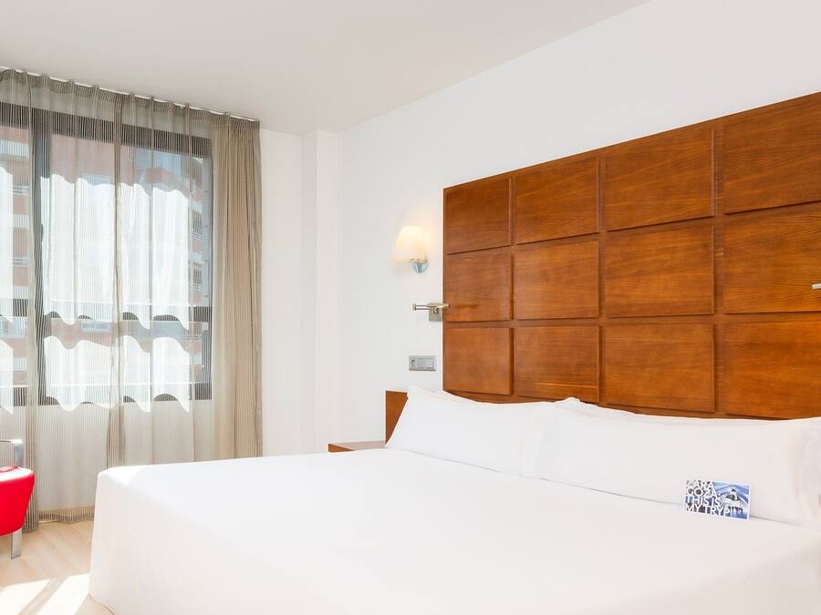 A hotel room with a large bed, a wooden headboard, a red chair, and sheer curtains.