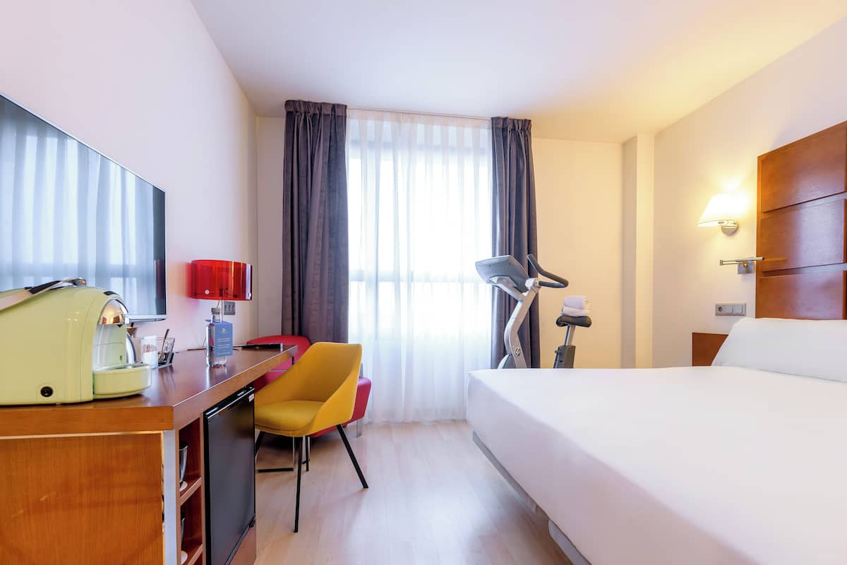 Habitación de hotel con cama, escritorio, silla, lámpara y cafetera.