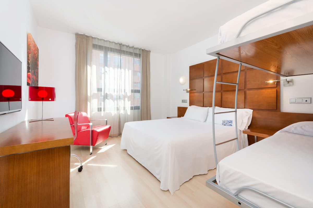 Una habitación de hotel con un escritorio de madera, una silla roja, dos camas, un televisor y un ventanal con cortinas.