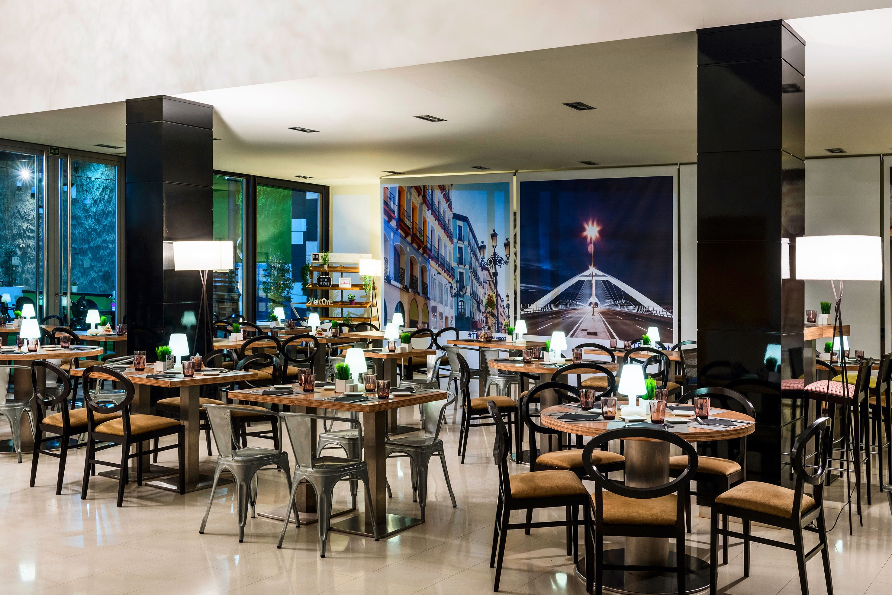 Un restaurante moderno con varias mesas y sillas, ventanas amplias y decoración mural ornamental.