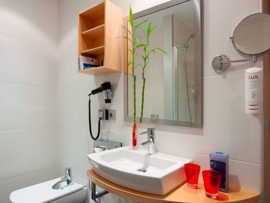 Un baño moderno con un lavamanos blanco, un espejo, una planta de bambú y un estante con artículos de aseo.