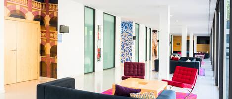 A modern hotel lobby with a seating area, a reception desk, and a view of the adjacent room.