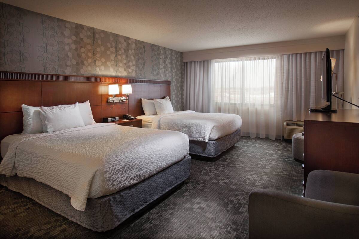 Une chambre d’hôtel avec deux lits, un bureau, une chaise, une lampe et une grande fenêtre avec des rideaux.