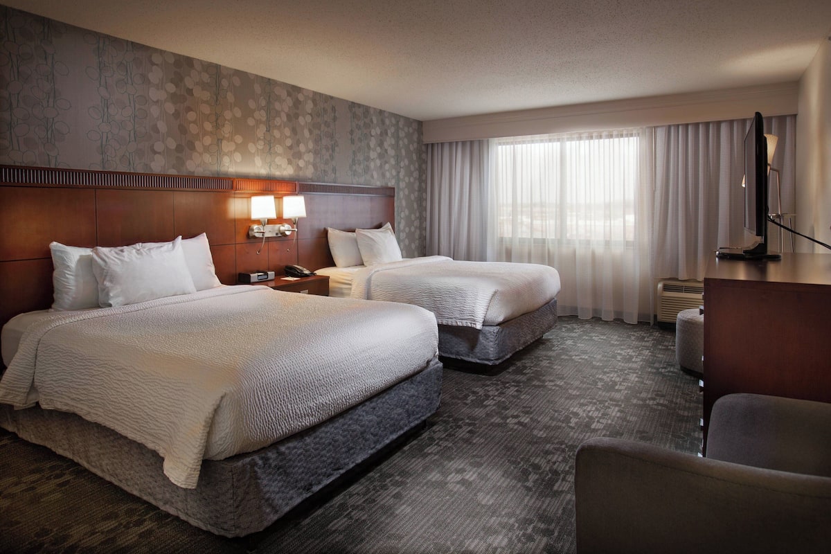 A hotel room with two beds, a desk, a chair, a lamp, and a large window with curtains.