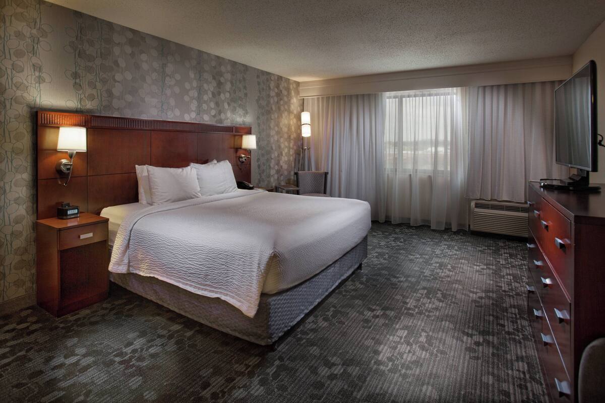Une chambre d’hôtel avec un grand lit, deux tables de chevet avec des lampes, un téléviseur à écran plat, une commode et une grande fenêtre avec des rideaux.