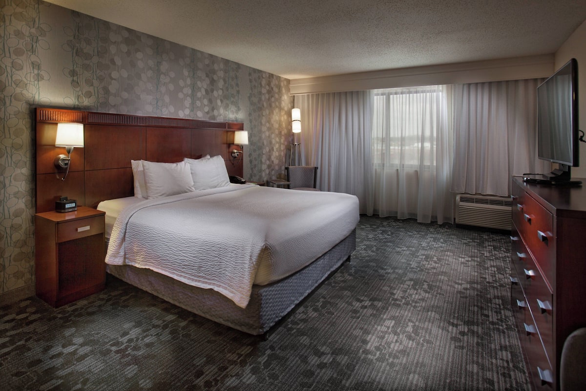 A hotel room with a large bed, two bedside tables with lamps, a flat-screen TV, a dresser, and a large window with curtains.