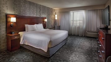 Una habitación de hotel con una cama grande, dos mesitas de noche con lámparas, un televisor de pantalla plana, un aparador y un ventanal con cortinas.
