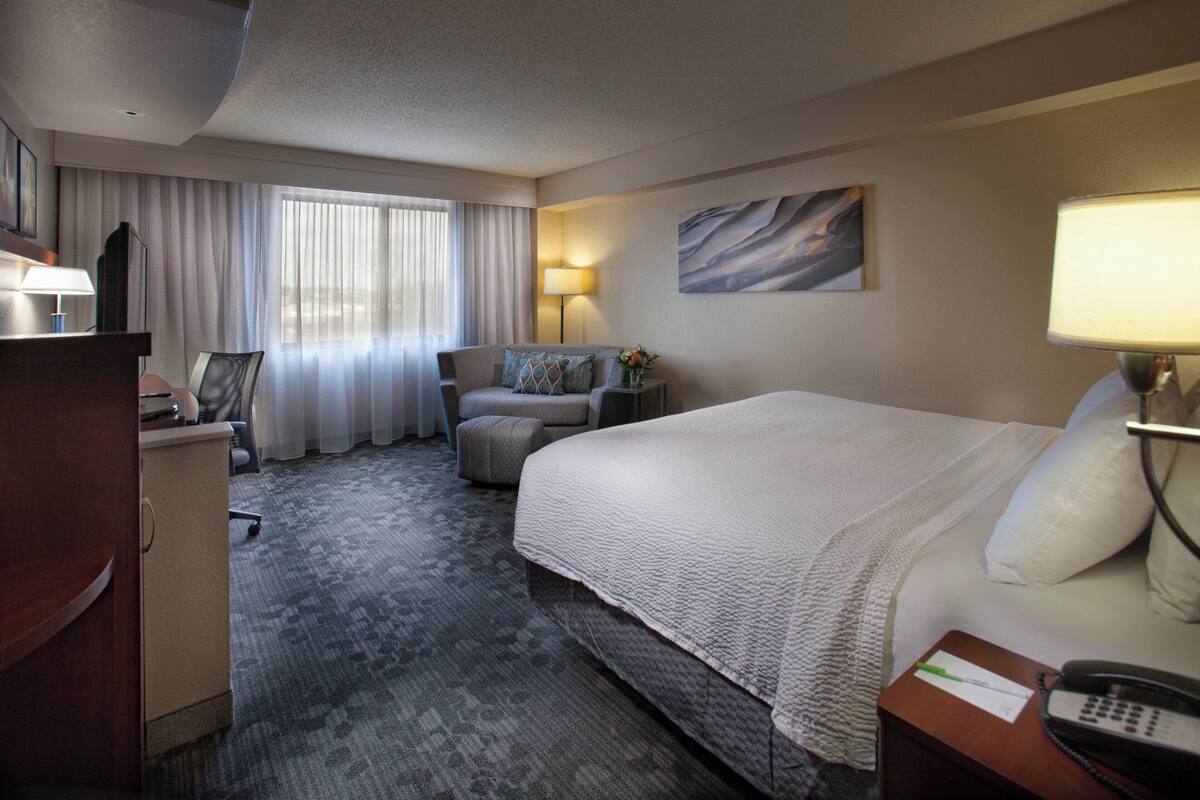 Une chambre d’hôtel avec un grand lit, un bureau avec une lampe, une chaise, une petite table avec un vase et un tableau au mur.