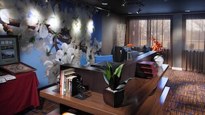 Un mostrador de recepción con una computadora, una silla azul y una planta. Se observa una pared con un mural floral y una ventana con cortinas.