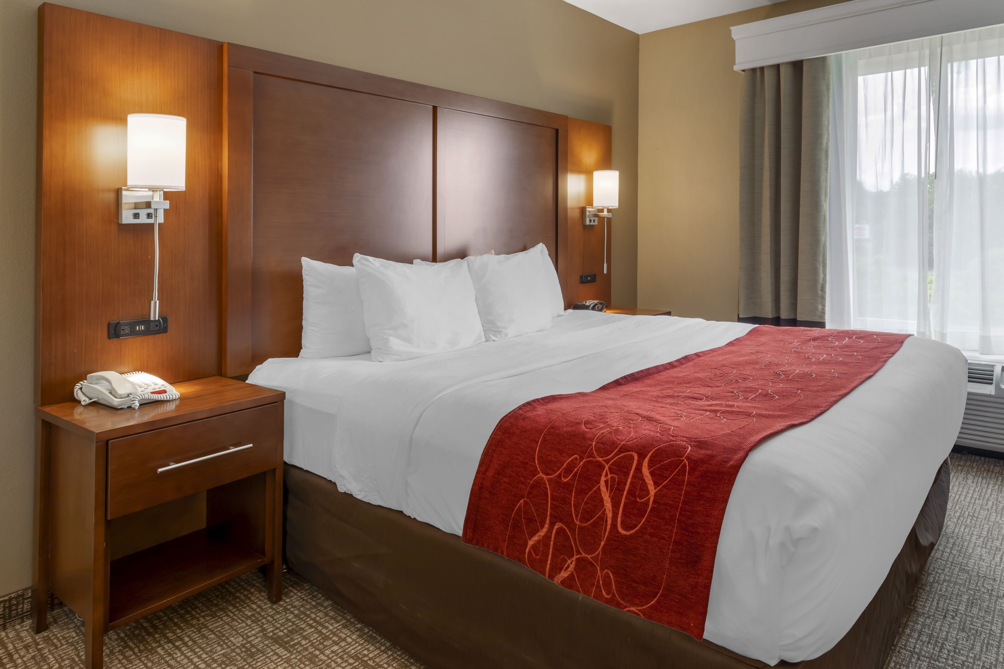 A hotel room with a large bed, wooden headboard, bedside lamps, a telephone, and a window with curtains.