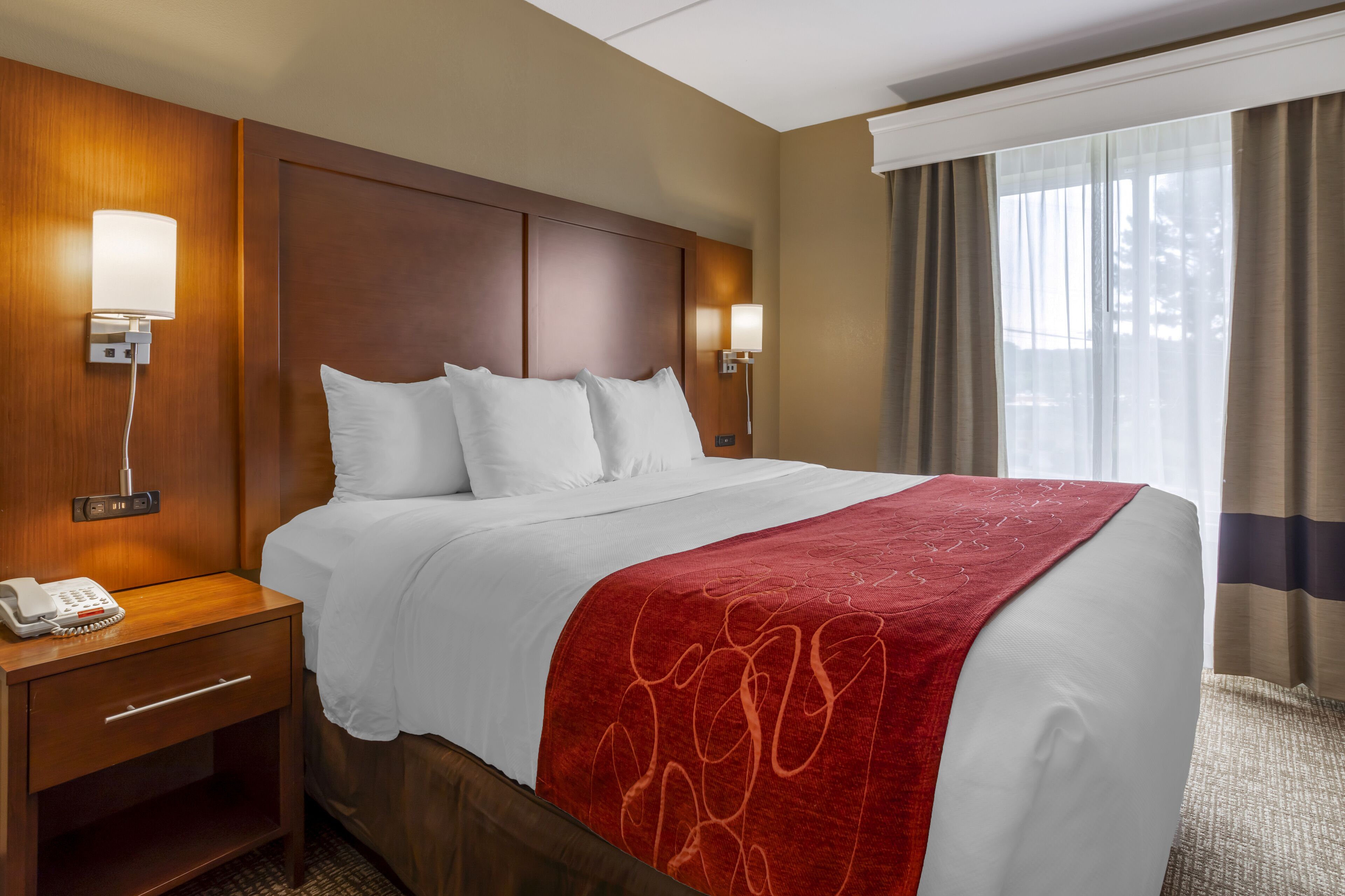 A hotel room with a large bed, a wooden nightstand with a phone, two wall-mounted lamps, and a window with curtains.