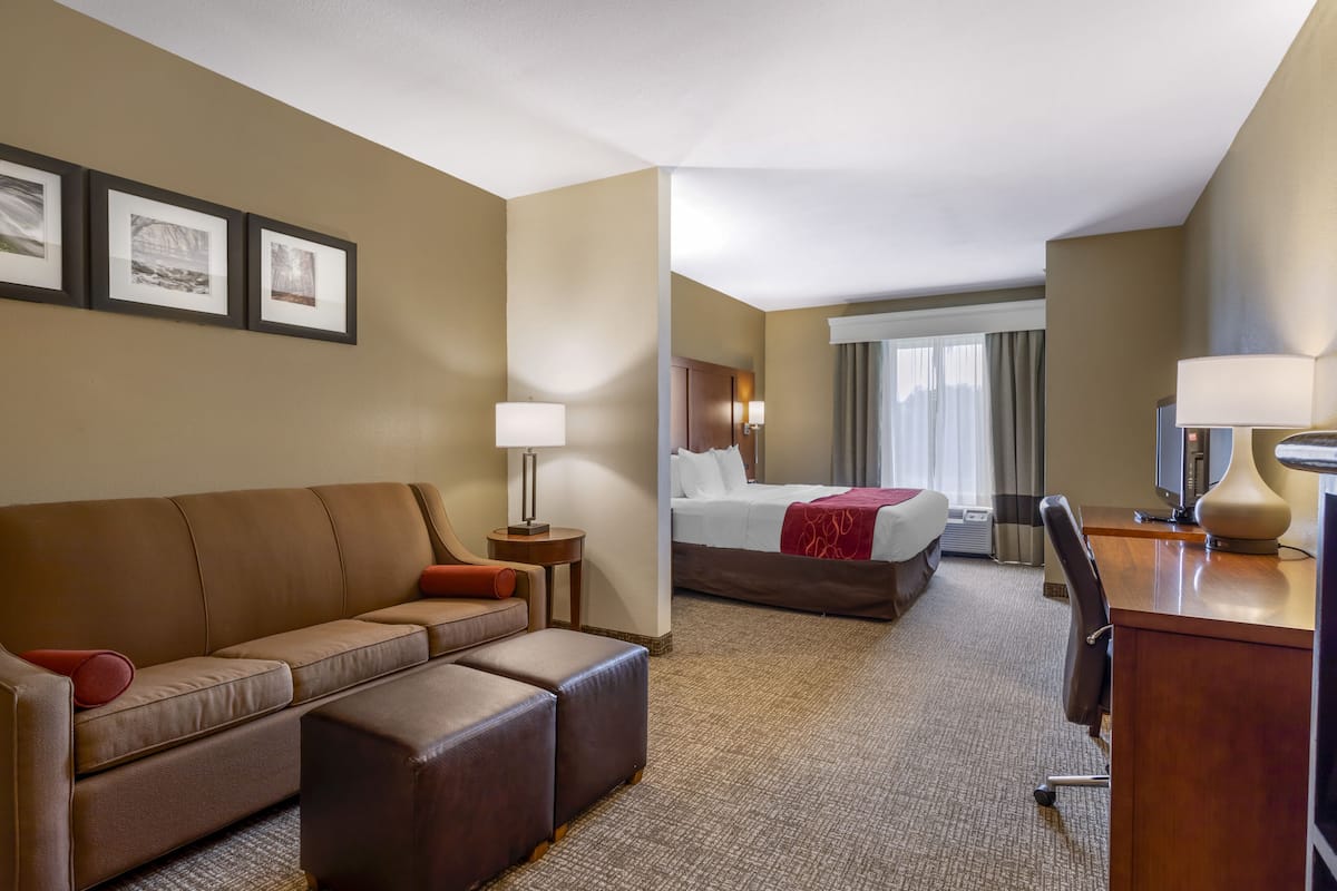 A hotel room with a brown leather sofa, a bed with a red blanket, a desk with a lamp, and framed pictures on the wall.