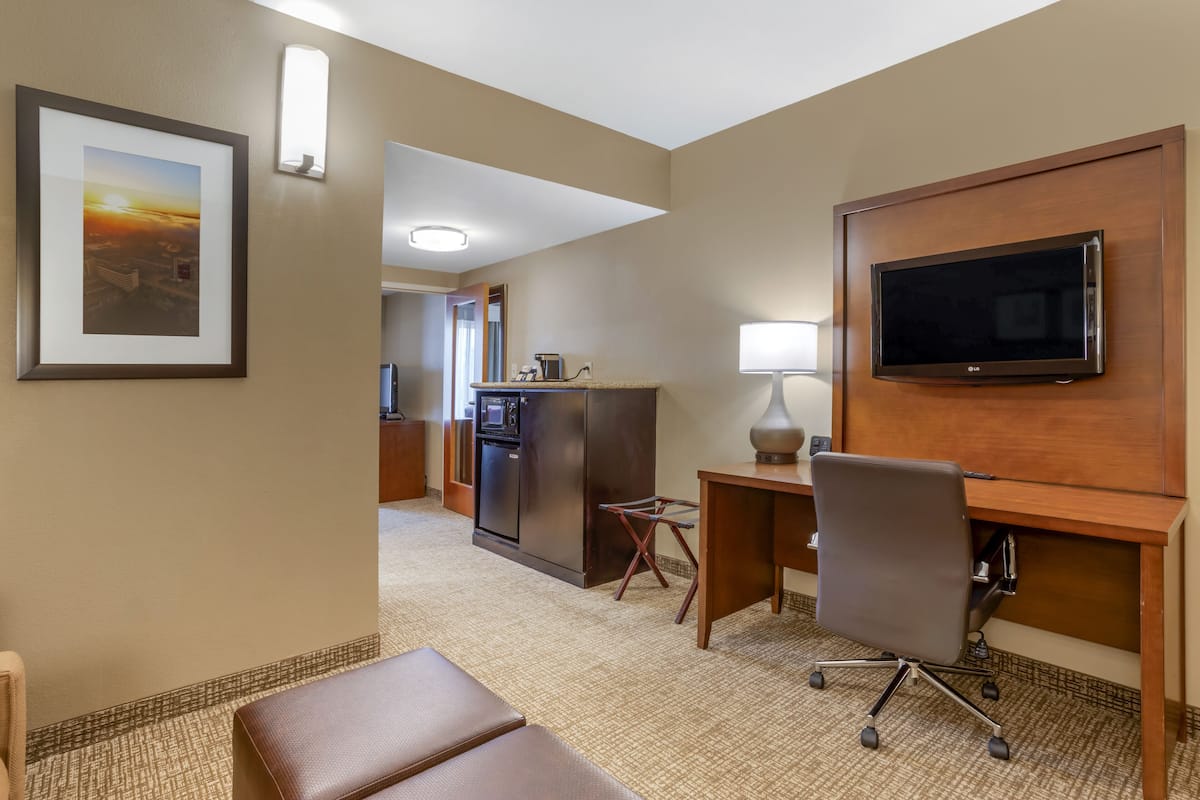 A hotel room with a desk, chair, TV, kitchenette, and a framed picture on the wall.