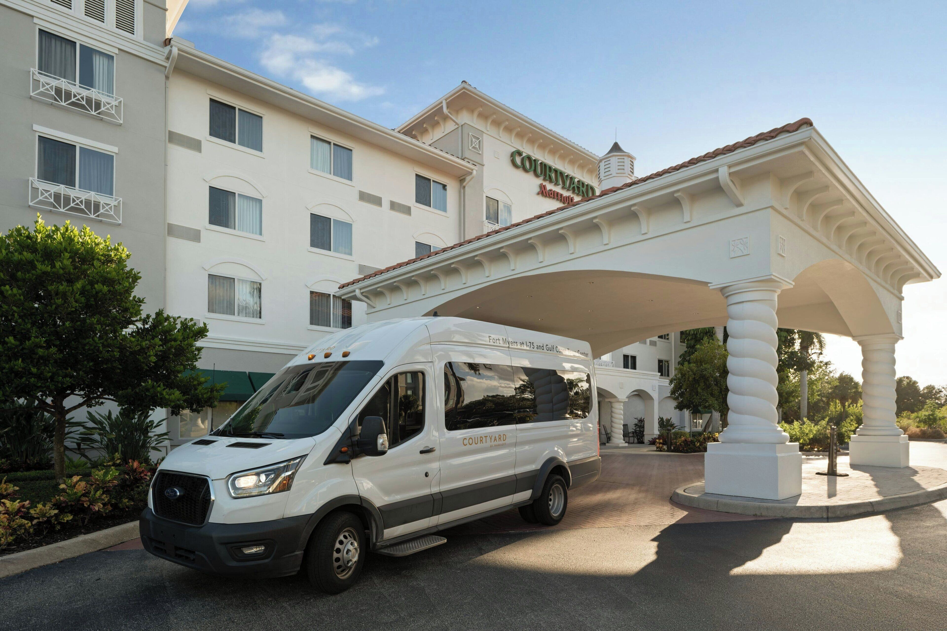 A white van parked under a white canopy with the text "COURTYARD" on it, in front of a hotel building with multiple windows and balconies.