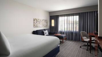 A hotel room with a large bed, a sofa, a desk, and a chair. There is a window with curtains and a painting on the wall.