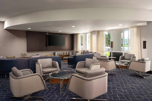 A modern hotel lobby with a curved ceiling, a large seating area with sofas and armchairs, a flat-screen TV mounted on the wall, and large windows allowing natural light.