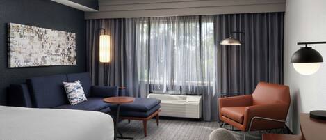 A modern hotel room with a bed, a sofa, an armchair, a small table, and a large window with curtains.
