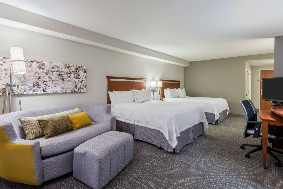 A hotel room with a gray sofa, two beds with white linens, a desk with a chair, and a large abstract painting on the wall.
