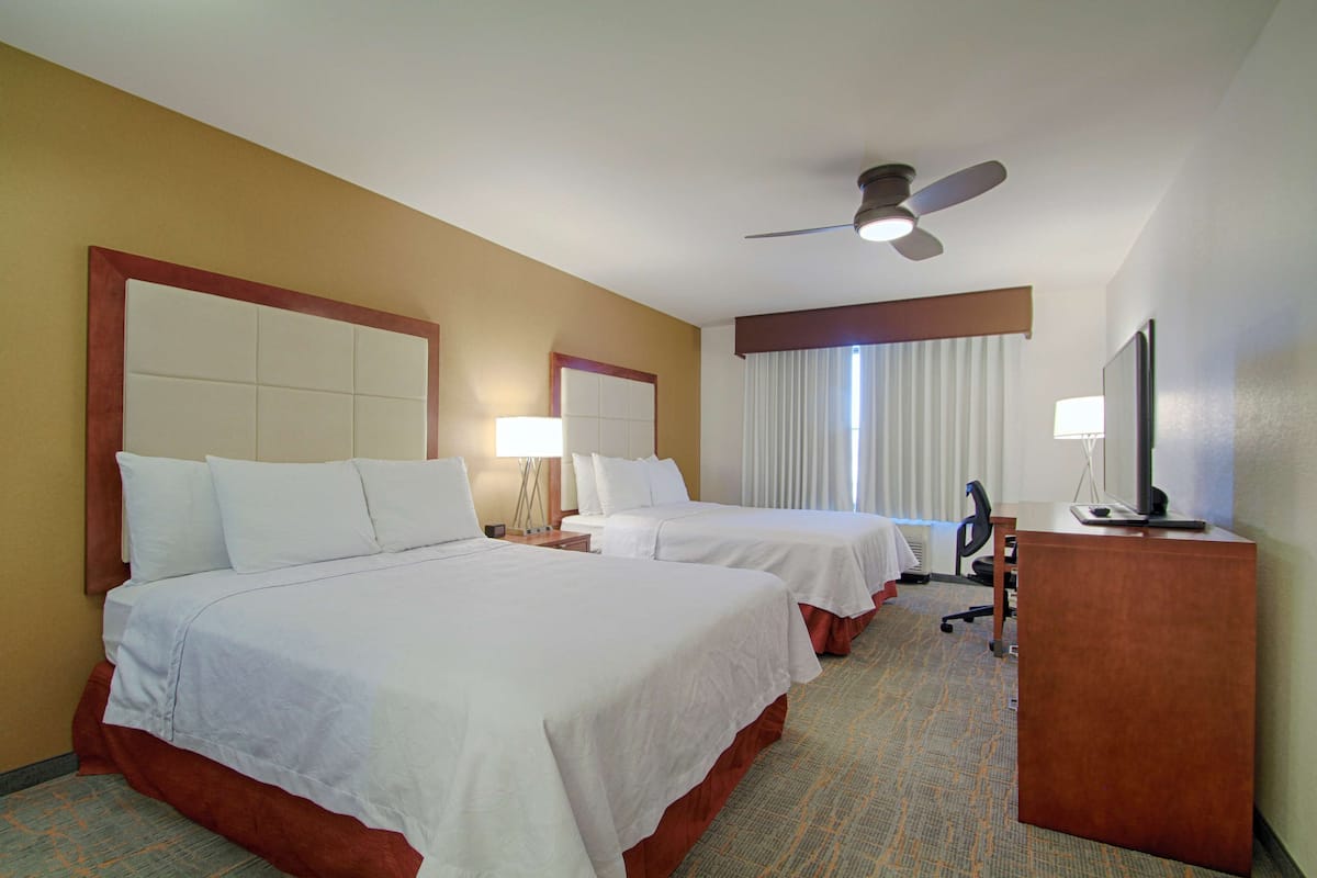 Ein Hotelzimmer mit zwei Betten, einem Schreibtisch, einem Sessel, einem Fernseher und einem Deckenventilator.