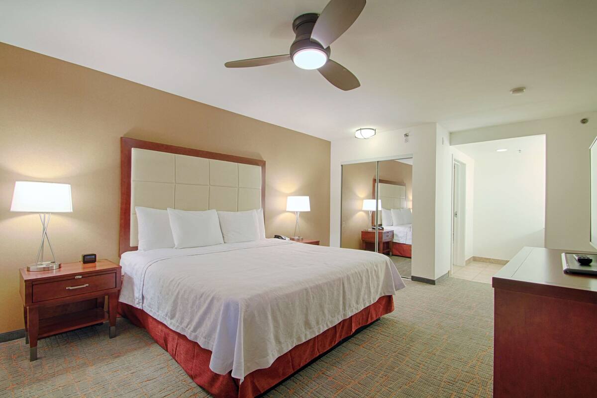 Ein Hotelzimmer mit einem großen Bett, einem Nachttisch, einem Schreibtisch, einem Deckenventilator und einem Spiegel.