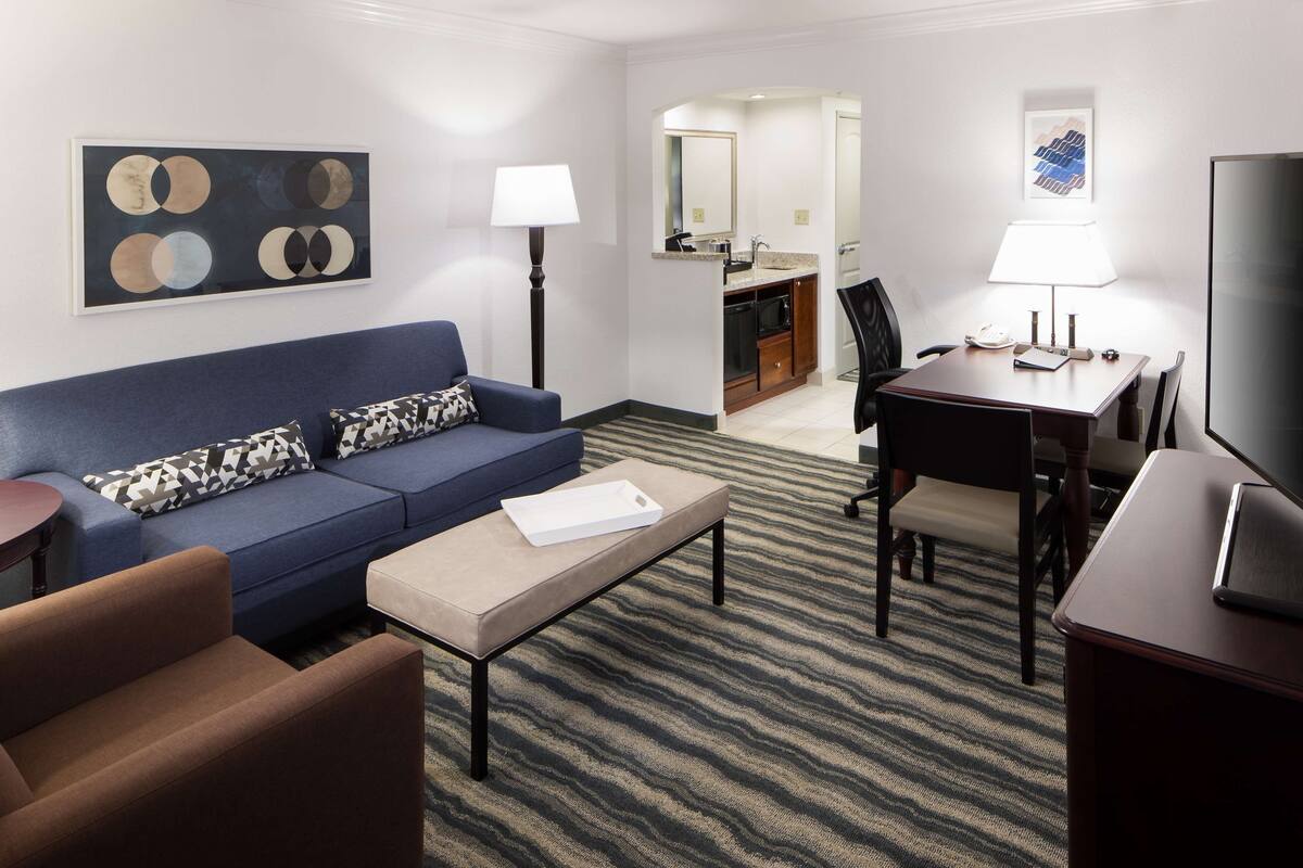 Une chambre d’hôtel moderne avec un canapé bleu, une petite table basse, une table à manger avec des chaises, un téléviseur et un coin cuisine à l’arrière-plan.
