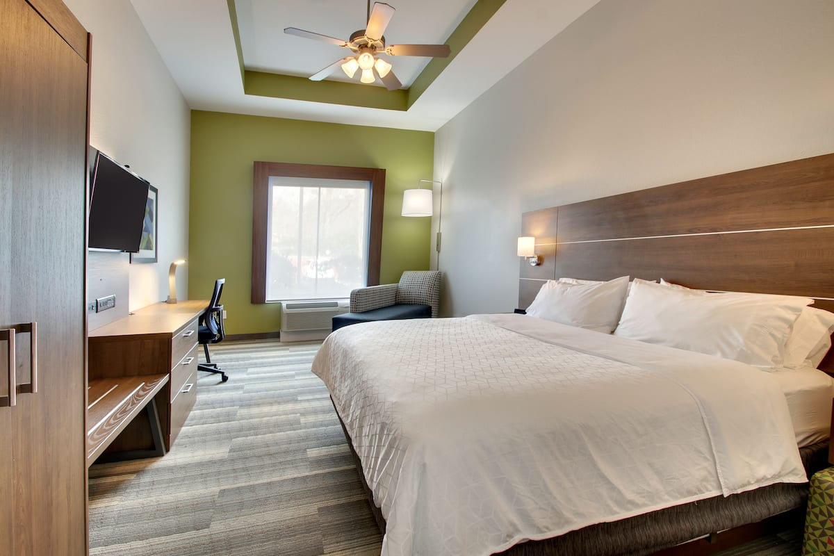 A hotel room with a large bed, a desk with a flat-screen TV, a ceiling fan, and a window with a view of the outdoors.