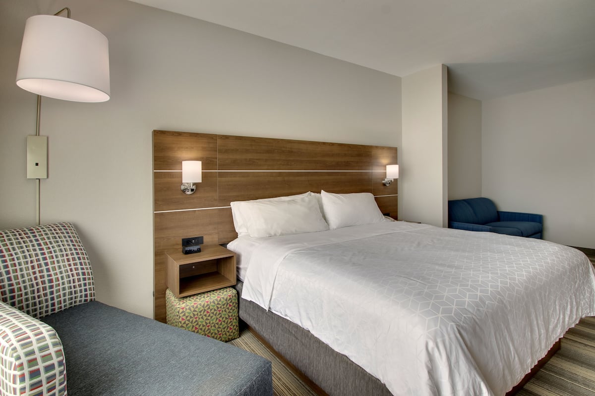 A hotel room with a large bed, a bedside table, a lamp, a chair, and a small ottoman.