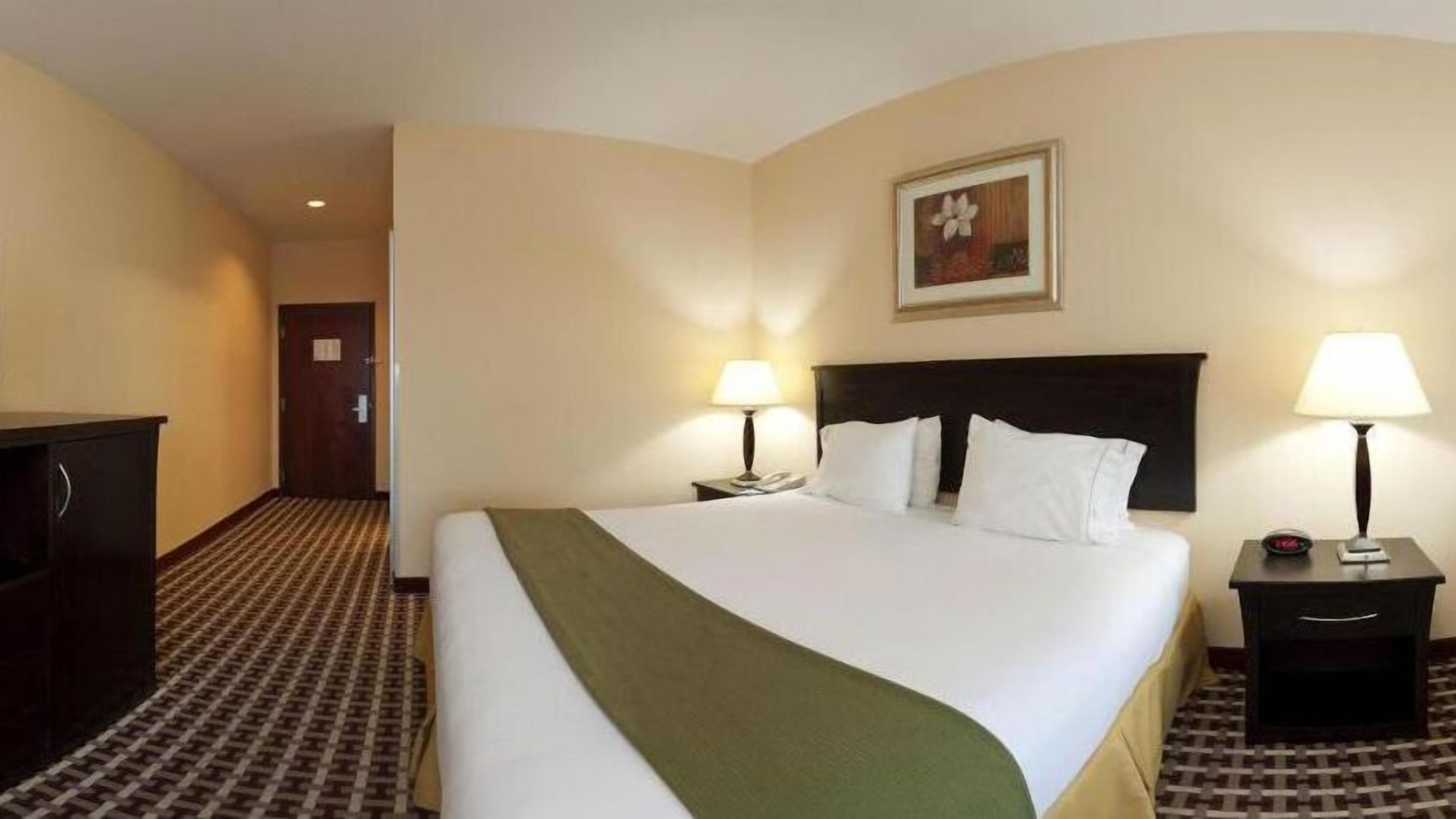 A hotel room with a large bed, two bedside lamps, a nightstand, a desk, and a framed picture on the wall.