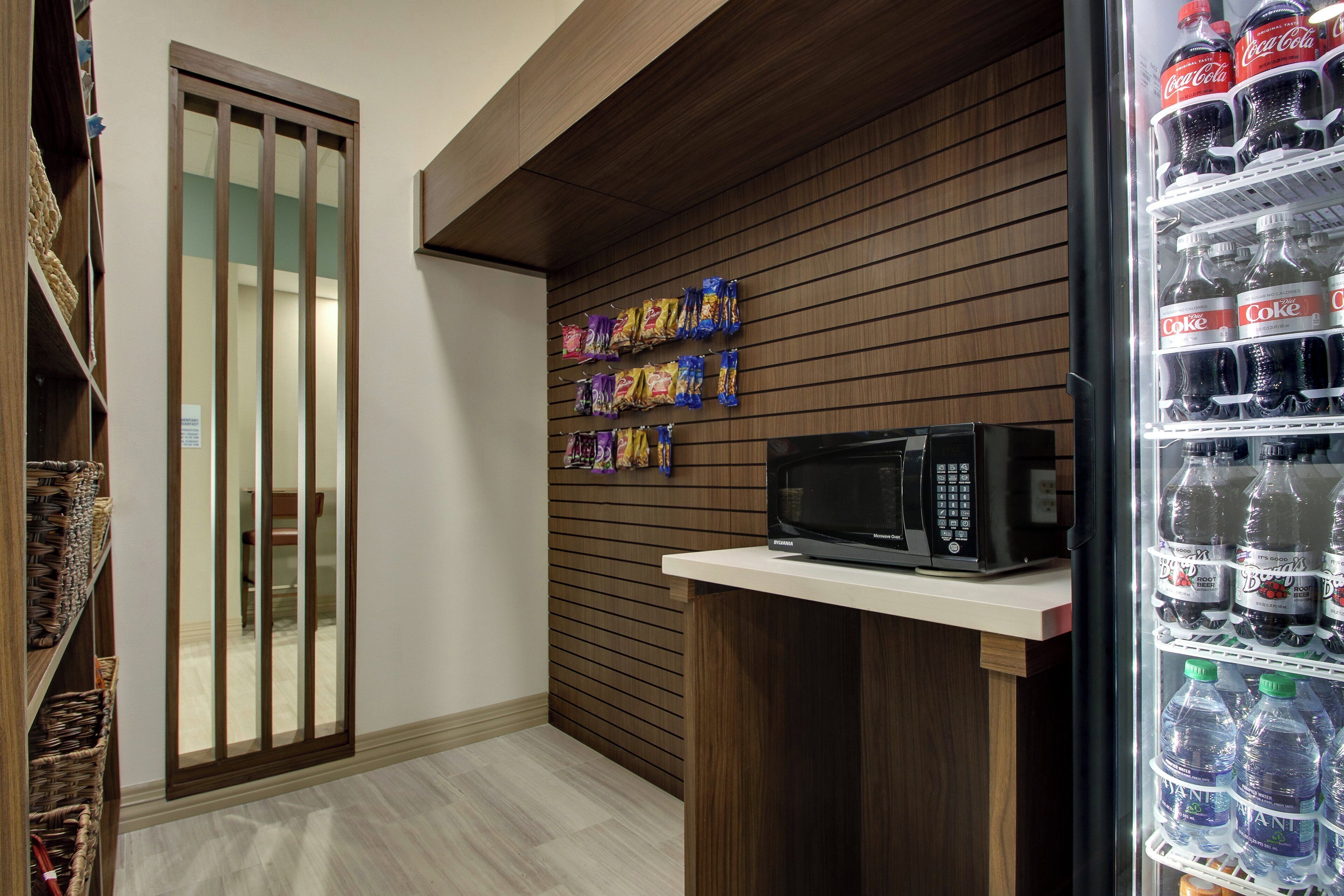 Un área de kitchenette con microondas, un refrigerador lleno de bebidas y paquetes de bocadillos en la pared.