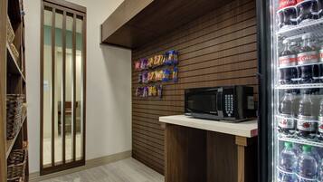 Un área de kitchenette con microondas, un refrigerador lleno de bebidas y paquetes de bocadillos en la pared.