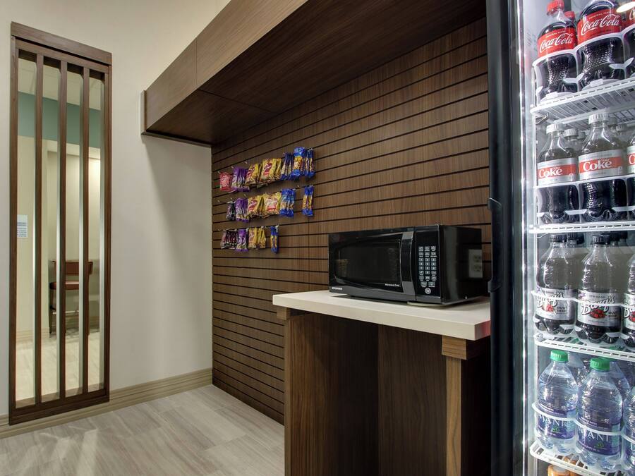 Un área de kitchenette con microondas, un refrigerador lleno de bebidas y paquetes de bocadillos en la pared.