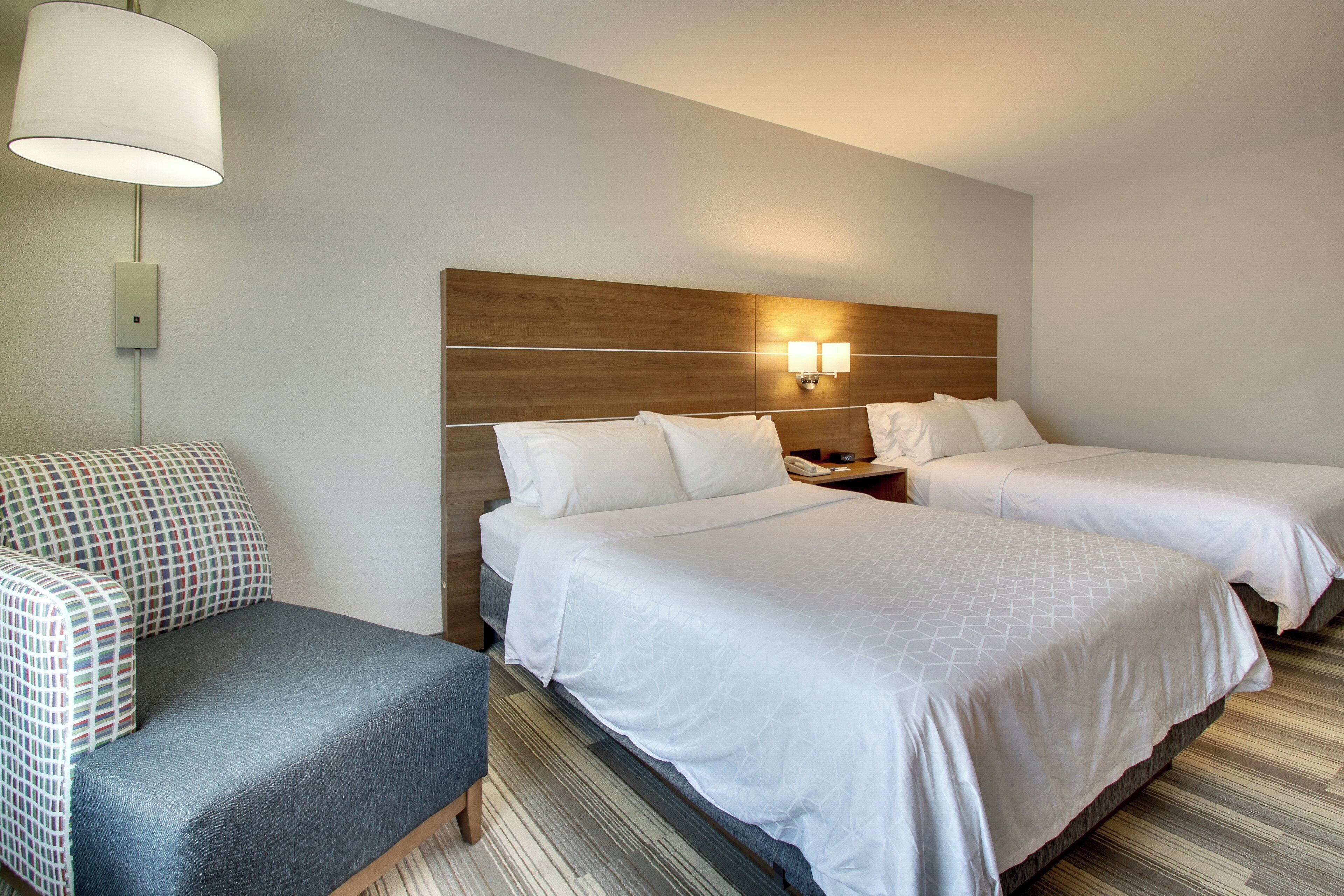 A hotel room with two beds, a chair, a nightstand, and a wall-mounted lamp.