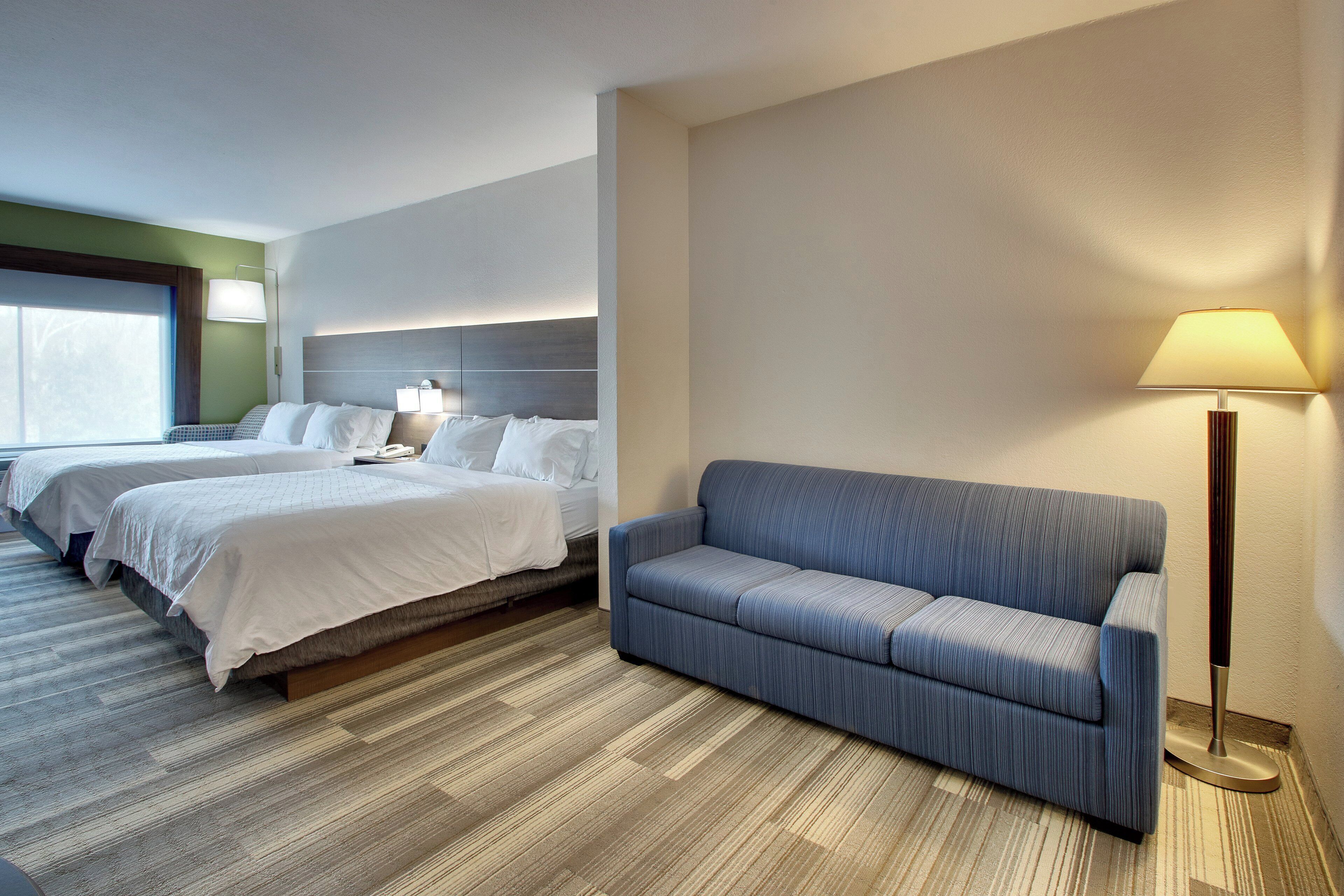 Quarto de hotel com duas camas, um sofá, uma luminária e uma janela grande.