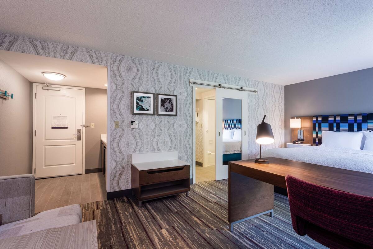 A hotel room with a bed, a desk, a chair, a small table, a nightstand, a lamp, and a bathroom with a sink and toilet.