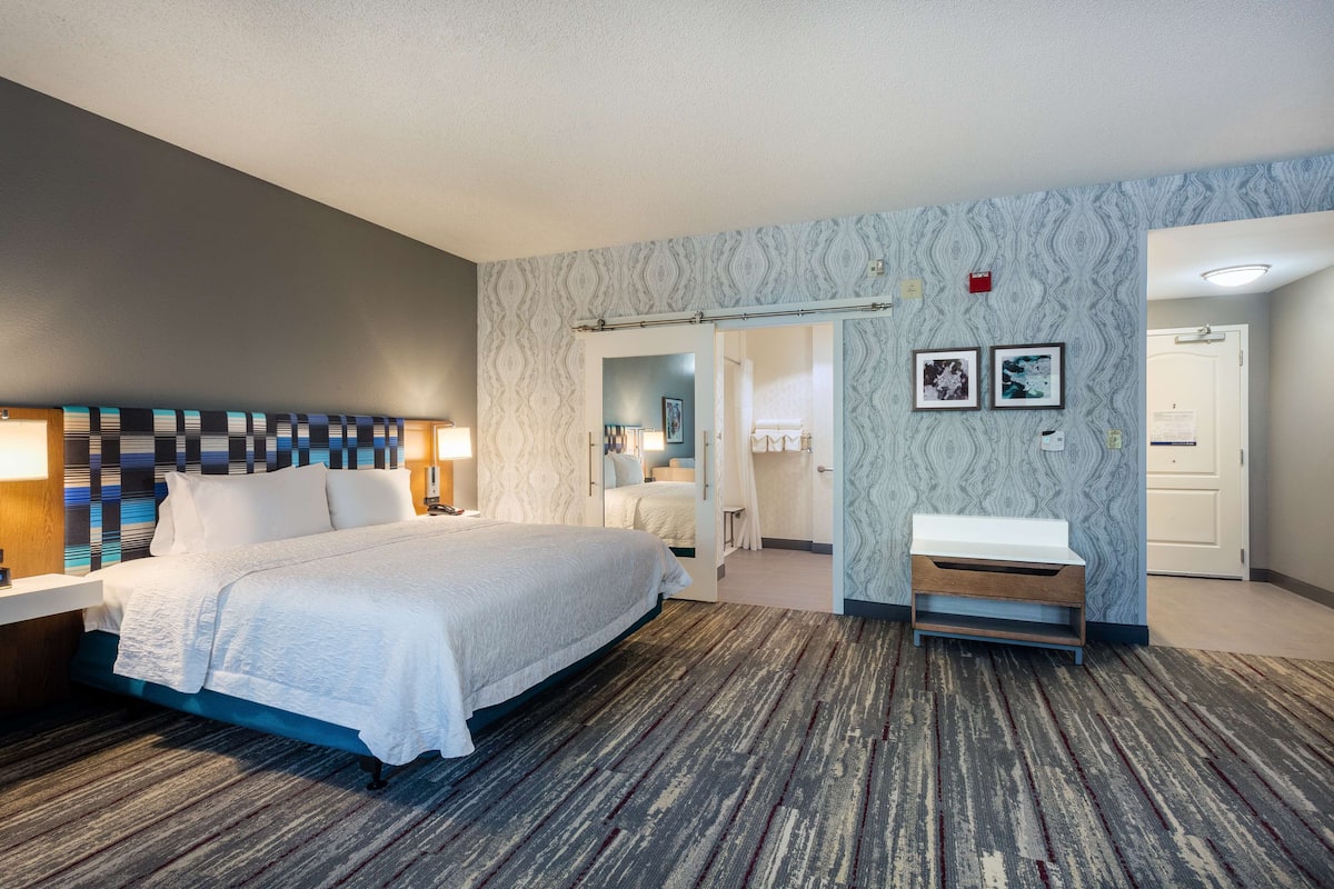 A hotel room with a large bed, a desk with a lamp, a vanity with a mirror, and a door leading to another room.