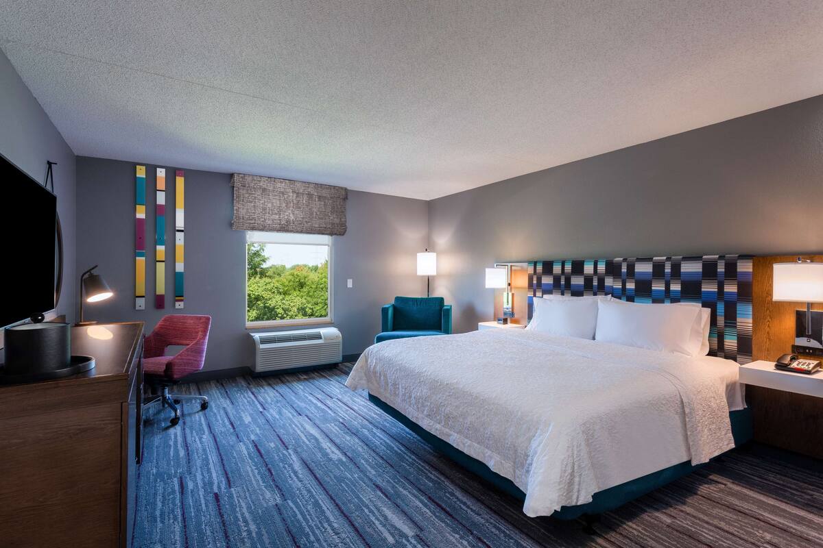 A modern hotel room with a large bed, a desk with a computer, a chair, a window with a view, and a patterned headboard.
