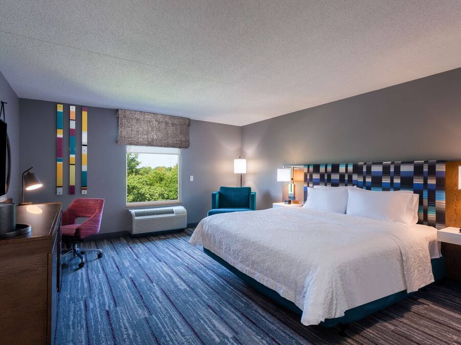Una habitación de hotel moderna con una cama grande, un escritorio con computadora, una silla, un ventanal con vista y un cabecero estampado.