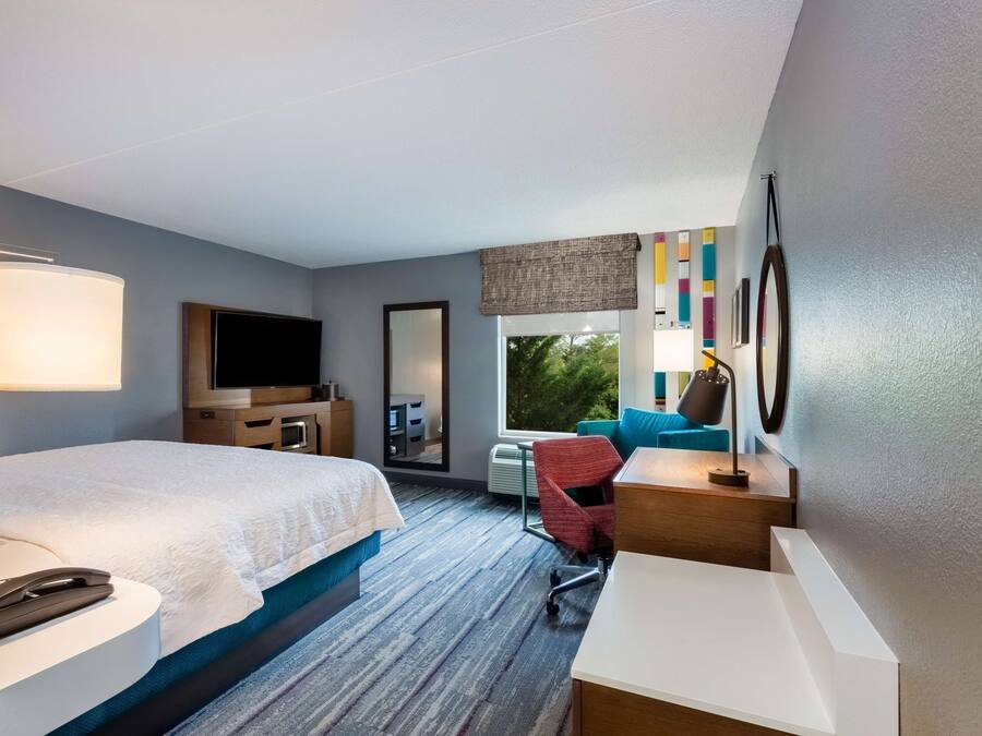A hotel room with a large bed, a desk, a chair, a TV, and a window with a view of greenery.