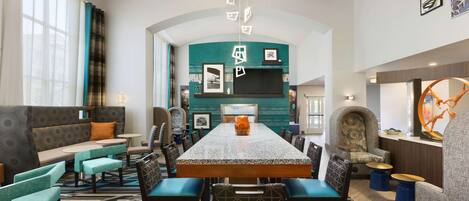 A modern hotel lobby with a long dining table, comfortable seating, and a large TV screen.