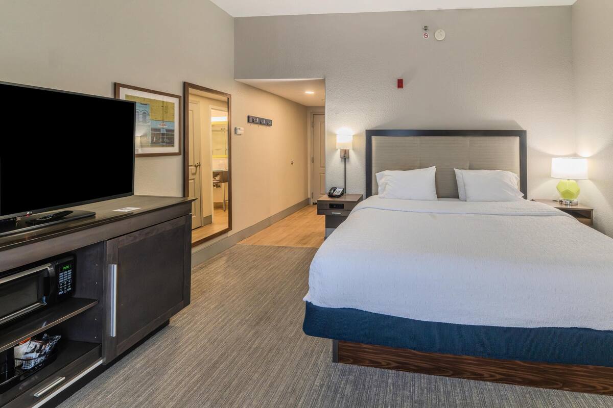 A hotel room with a bed, a TV, a minibar, and a bathroom door.