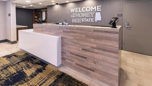 A reception desk with a "Welcome to the Honey Bee State" sign, a digital kiosk, and a reception area with a patterned carpet. - Hampton Inn Jasper (Jasper)