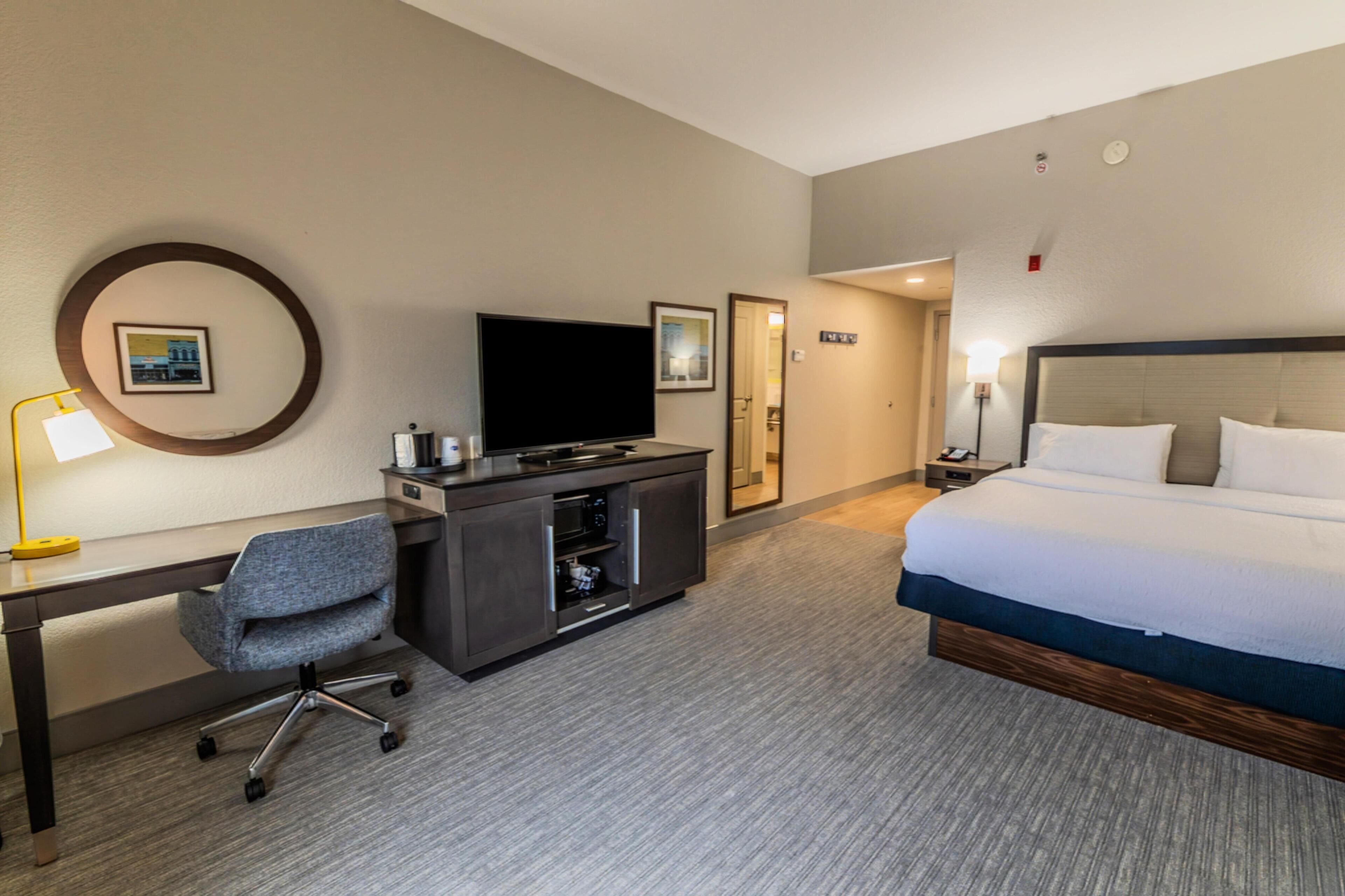 A hotel room with a bed, a desk with a computer, a TV, and a small kitchenette.