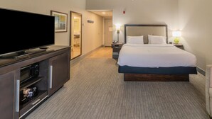 A hotel room with a large flat-screen TV, a bed with white linens, a wooden headboard, a nightstand with a lamp, and a built-in microwave. - Hampton Inn Jasper (Jasper)