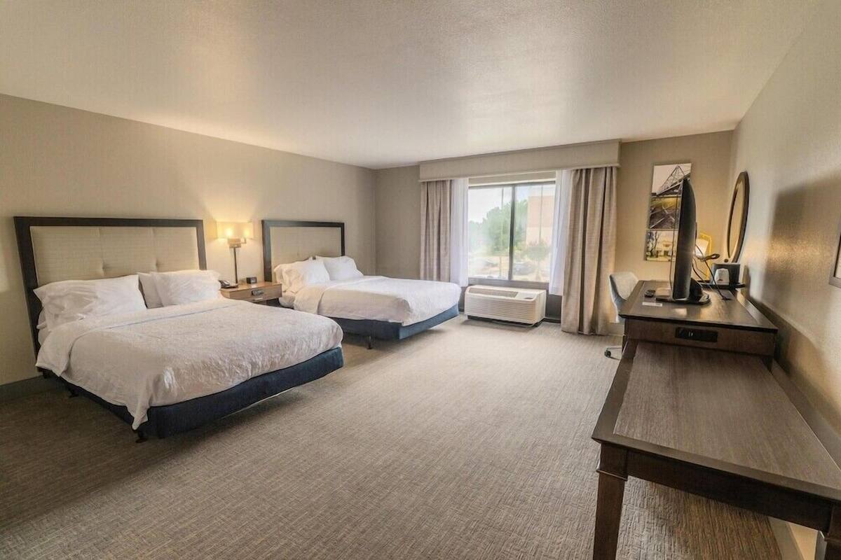 A hotel room with two beds, a desk, a chair, a lamp, and a window with curtains.