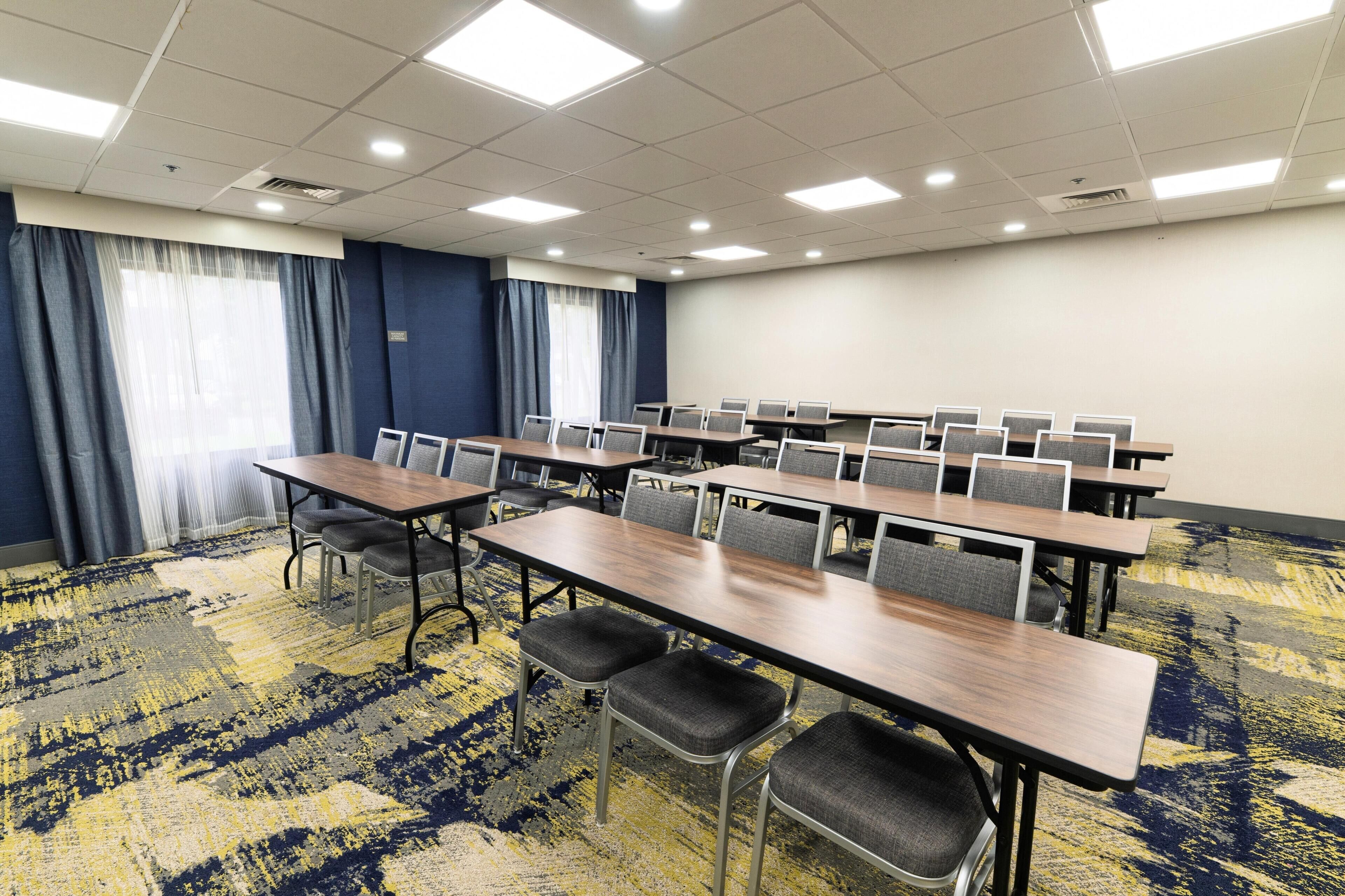 Un salón de conferencias con varias mesas y sillas, alfombra estampada y grandes ventanales con cortinas.